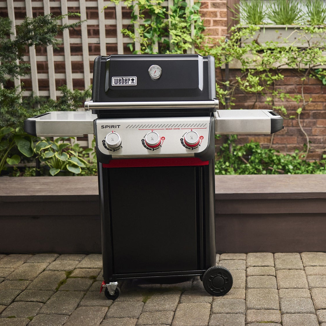 Black and red grill on a patio with greenery and a brick wall in the background