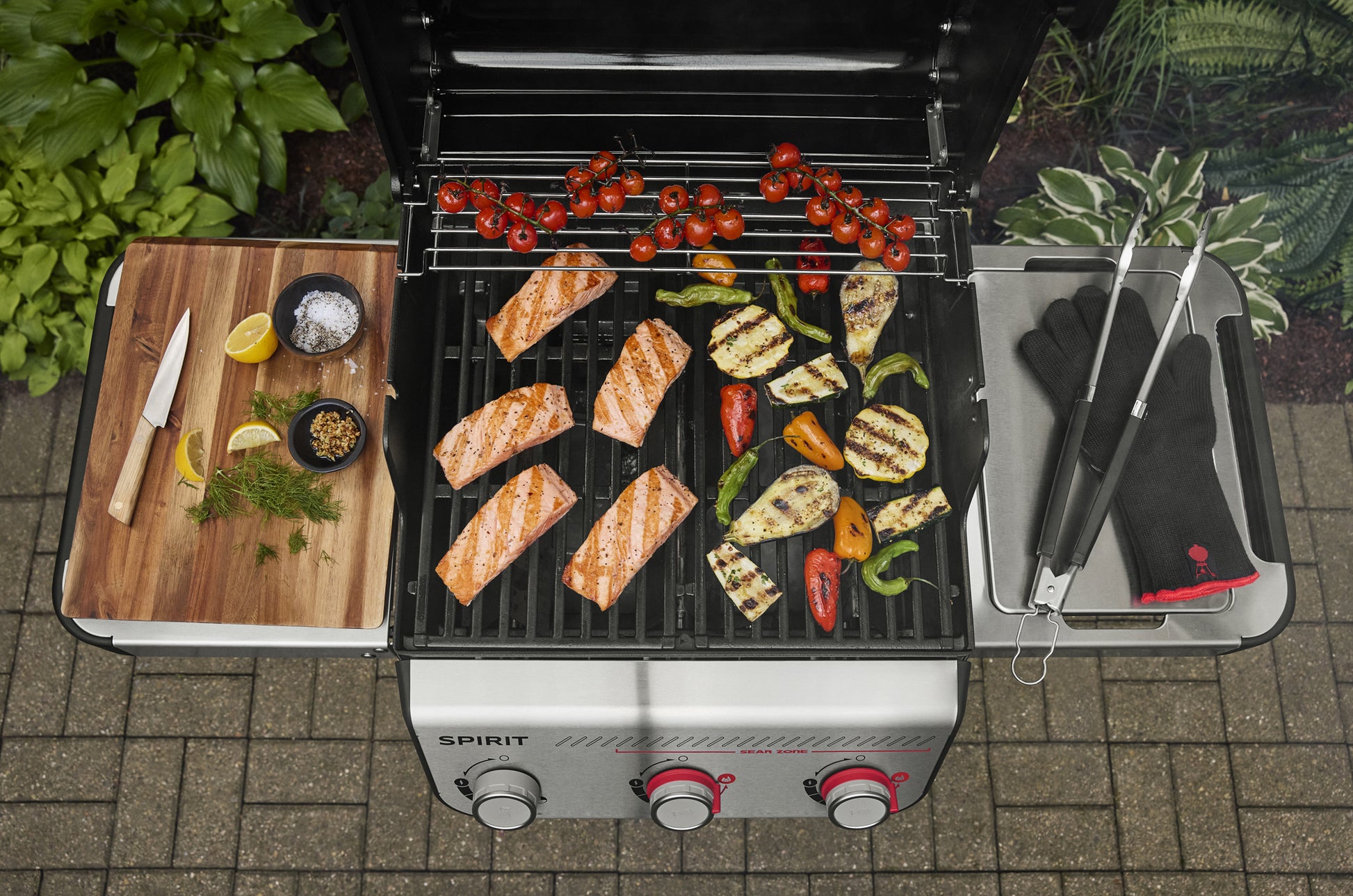 Outdoor grill with food being cooked, including salmon and vegetables, on a patio.