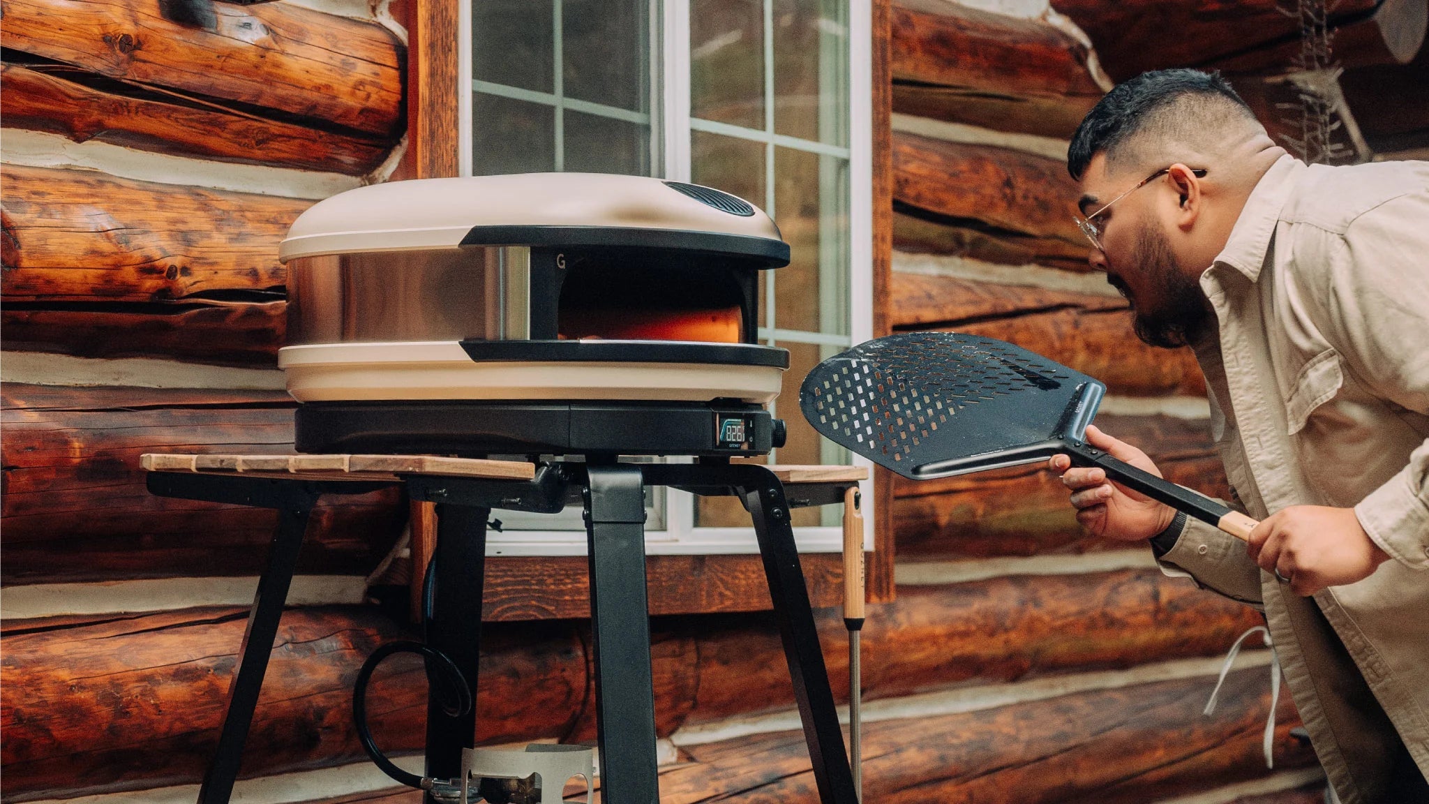 Person using a portable pizza oven on a wooden deck with a log cabin background